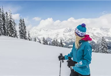 Eine Skifahrerin in einer blauen Jacke steht in einer verschneiten Landschaft. Im Hintergrund sind hohe Berge unter einem klaren Himmel zu sehen.