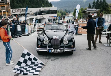 A classic car crosses the finish line in a race. Spectators and a black-and-white flag are visible in the background.