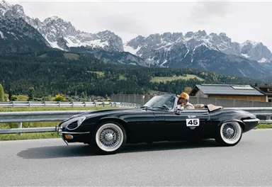 A black convertible drives along a winding road with impressive mountains in the background. The driver is wearing a hat and enjoying the ride in nature.
