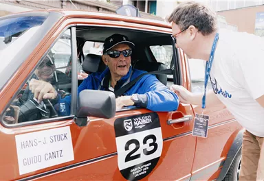 An older man in the car is talking to a younger man. The car has a racing number and a sticker with the names of the drivers.