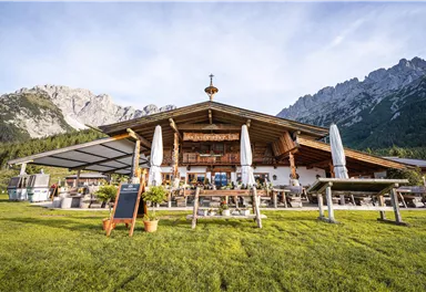 Ein traditionelles Alpenhaus mit großzügiger Terrasse und Sonnenschirmen. Im Hintergrund erheben sich majestätische Berge unter einem klaren Himmel.