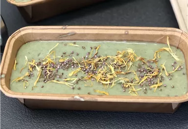 Ein gefüllter Behälter mit einer grünen, cremigen Masse. Die Oberfläche ist mit bunten Samen und getrockneten Blüten dekoriert.