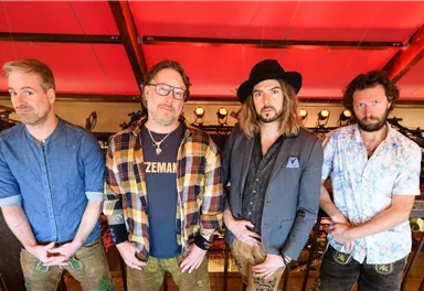 Eine Gruppe von vier Männern steht auf einer Tribüne. Sie tragen verschiedene Outfits und posieren vor einem roten Hintergrund.