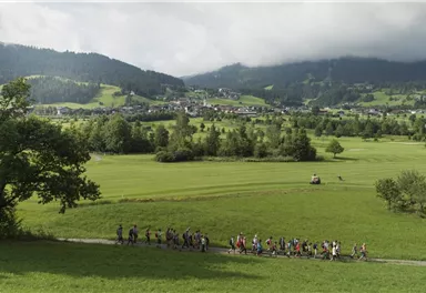 Eine Gruppe von Menschen wandert auf einem Weg durch eine grüne Landschaft. Im Hintergrund sind Hügel und ein kleiner Ort zu sehen.
