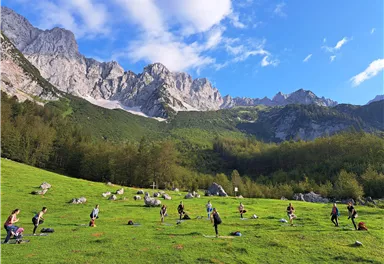 Eine Gruppe von Menschen trainiert in einer grünen Wiese mit Bergen im Hintergrund. Der Himmel ist blau mit wenigen Wolken.