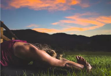 Eine Person macht Yoga im Gras mit einer wunderschönen Berglandschaft im Hintergrund. Der Himmel ist bei Sonnenuntergang in sanfte Farben getaucht.