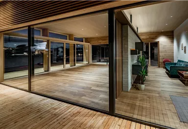A modern, light-filled living room with wooden flooring and large windows. It has a minimalist decor and a view of the surroundings.
