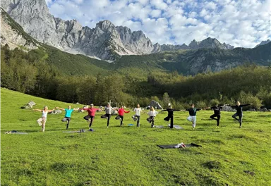 Eine Gruppe von Menschen praktiziert Yoga auf einer Wiese in den Bergen. Die Landschaft ist grün mit majestätischen Felsen im Hintergrund und einem blauen Himmel.