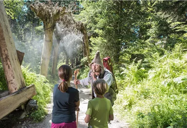 Ein Wanderer im Wald erzählt Kindern Geschichten. Die Umgebung ist grün und idyllisch, mit vielen Pflanzen und Bäumen.