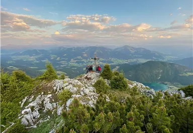 Eine beeindruckende Berglandschaft mit Aussicht auf grüne Täler und Berge. Im Vordergrund steht ein Gipfel mit Menschen und einem Kreuz.