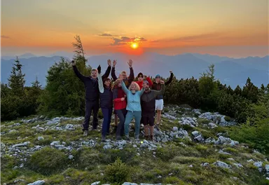 Eine Gruppe von Menschen steht auf einem Berg und jubelt beim Sonnenuntergang. Die Landschaft ist mit Bäumen und Felsen umgeben.