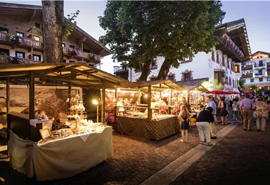 Ein lebendiger Marktplatz bei Dämmerung mit Ständen, die lokale Produkte anbieten. Menschen schlendern durch die Gassen und genießen die Atmosphäre.