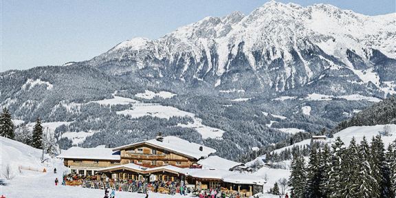 Eine malerische Winterlandschaft mit schneebedeckten Bergen. Im Vordergrund steht eine Berghütte, umgeben von Skifahrern und Tannenbäumen.