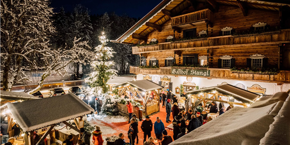 Eine festliche Wintermarkt-Szene mit Menschen, die Stände besuchen. Im Hintergrund steht ein rustikales Holzhaus, umgeben von schneebedeckten Bäumen und einem beleuchteten Weihnachtsbaum.