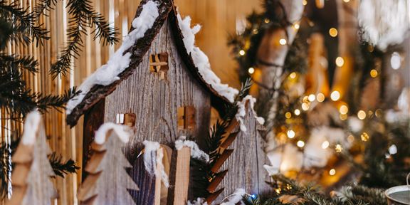 Ein dekoratives Holzhaus mit Schnee und kleinen Tannenbäumen. Im Hintergrund sind Lichter und festliche Dekorationen zu sehen.