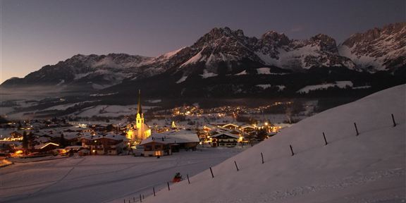 A picturesque alpine landscape at night with snow-covered mountains. In the foreground, a small village with illuminated houses and a church is visible.