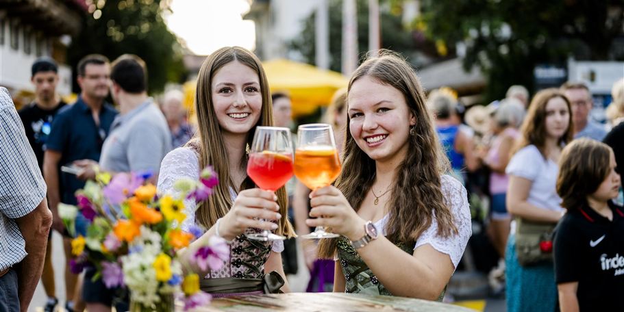 Zwei junge Frauen stoßen mit bunten Getränken an. Im Hintergrund sind Menschen und Marktstände zu sehen, die eine festliche Atmosphäre erzeugen.
