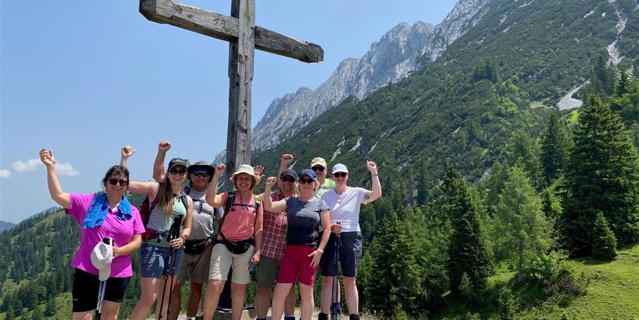 A group of hikers stands in front of a large wooden cross in the mountains. They are smiling and happily posing on a sunny day.