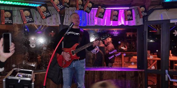 Ein Musiker mit einer roten Gitarre steht auf einer kleinen Bühne und singt. Im Hintergrund sind bunte Lichter und festliche Dekorationen zu sehen.