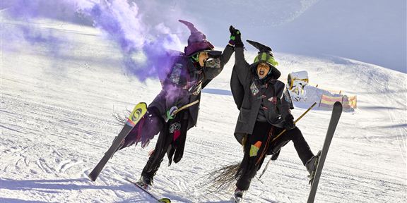 Two costumed witches are celebrating on the ski slope, holding hands. Colorful smoke flares surround them in the snowy landscape.