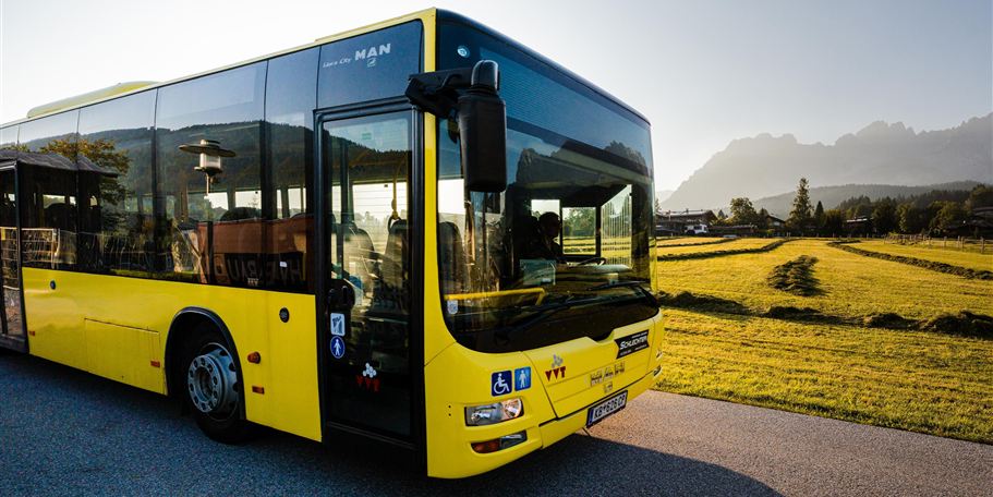 Ein gelber Bus steht auf einer Straße neben einem Feld. Im Hintergrund sind Berge und ein klarer Himmel zu sehen.
