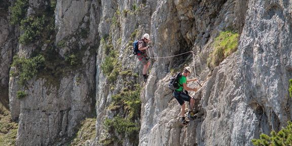 Two climbers scaling a steep rock face. The surroundings are green and mountainous, with plenty of natural light.