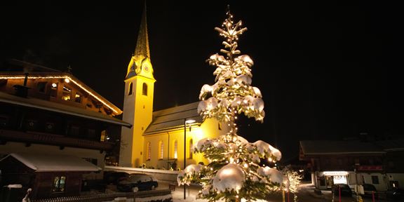 Ein festlich geschmückter Weihnachtsbaum steht im Schnee vor einer beleuchteten Kirche. Die Szenerie ist nachts und strahlt eine gemütliche Stimmung aus.