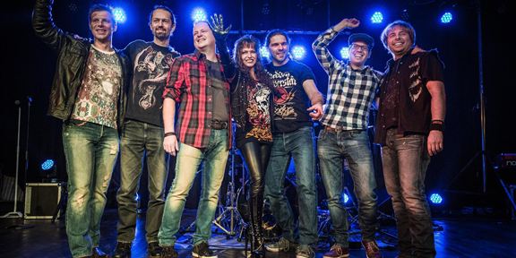 Eine Gruppe von sieben Musikern steht auf einer Bühne. Sie lächeln und posieren vor einem festlichen Hintergrund mit blauen Lichtern.