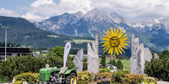 Ein grüner Traktor steht vor einem sonnigen, blühenden Blumenbeet. Im Hintergrund sind majestätische Berge und ein strahlend blauer Himmel zu sehen.