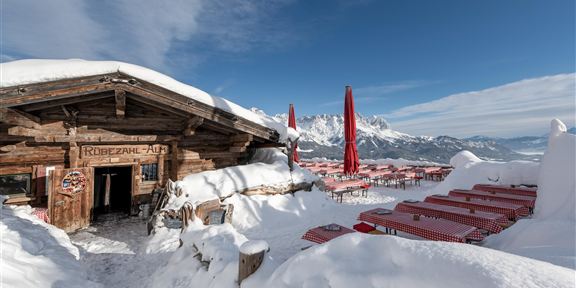 Eine rustikale Berghütte in einer verschneiten Berglandschaft. Rote Sonnenschirme stehen an den Tischen im Freien.