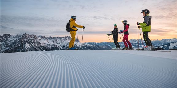 A group of skiers stands on a freshly groomed slope in the mountains. The view shows snow-covered peaks and a clear sky at sunrise.
