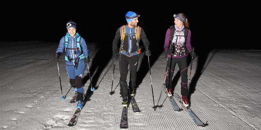 Drei Skifahrer gehen nachts über einen schneebedeckten Hang. Sie tragen Stirnlampen und skifahren in einem tiefen, dunklen Umfeld.