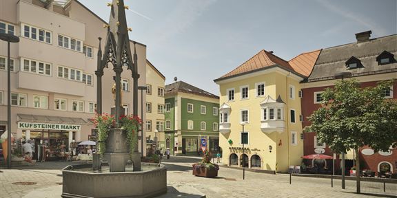 Ein schöner Platz mit einem Brunnen in der Mitte und bunten Gebäuden im Hintergrund. Die Atmosphäre ist ruhig und einladend.