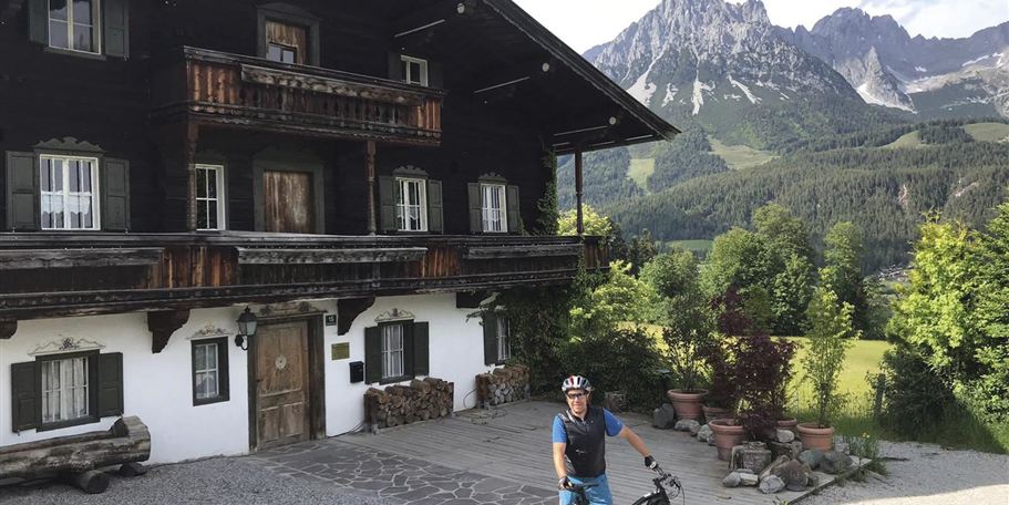 A cyclist stands in front of a traditional chalet in the mountains. In the background, green meadows and impressive mountain peaks can be seen.