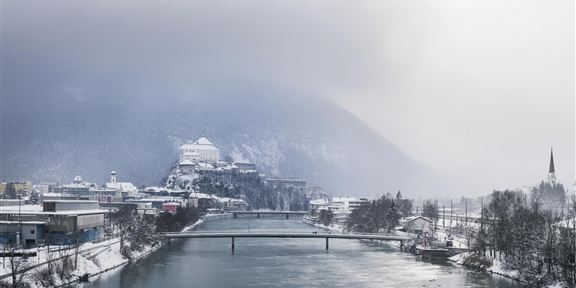 Eine verschneite Stadt am Ufer eines Flusses. Im Hintergrund sind Berge und grauer Himmel zu sehen.