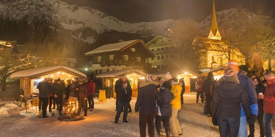 A wintry Christmas market at night with festively illuminated stalls. People enjoy the atmosphere in a snow-covered setting, surrounded by mountains.