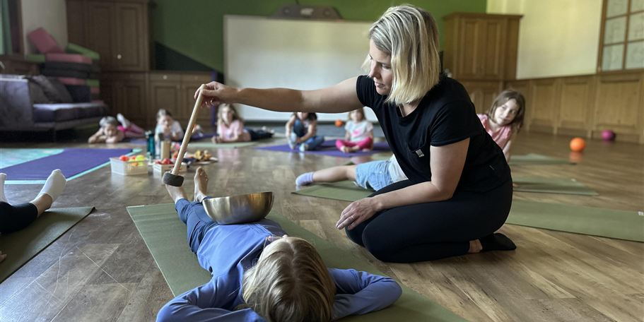 A teacher conducts a relaxation exercise with children. The children are lying on mats while the teacher works with a bowl and a spoon.