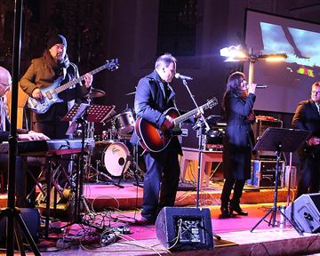 A band is playing live on a stage with various musical instruments. In the background, lights and a screen can be seen.