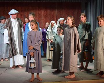 A group of children and adults in historical costumes stands on a stage. They are holding lanterns and are attentively looking towards the audience.