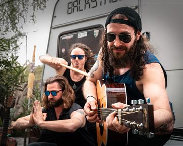 Three musicians are playing in a relaxed atmosphere. In the background, plants and a caravan can be seen.