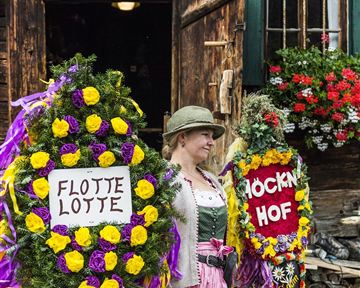 Eine Frau in traditioneller Tracht steht vor einem Holzhaus und trägt zwei festlich geschmückte Kränze. Die Kränze sind mit bunten Blumen dekoriert und tragen die Schriftzüge "FLOTTEN LOTTE" und "HÖCKEN HOF".