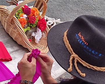 Eine Person bastelt Blumen aus buntem Stoff. Im Hintergrund ist ein Korb mit bereits fertiggestellten Blumen zu sehen.