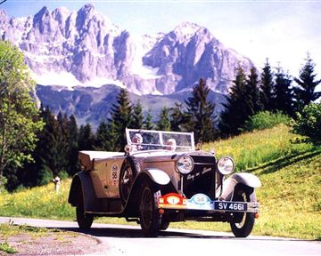 Ein klassisches Auto fährt eine malerische Straße in den Bergen. Die Umgebung ist grün mit hohen Bäumen und beeindruckenden Berggipfeln im Hintergrund.