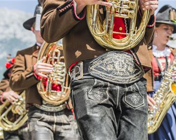 A group of musicians in traditional clothing is playing wind instruments. In the background, mountains and a sunny landscape can be seen.
