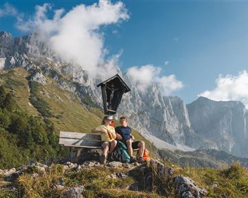 Zwei Personen sitzen auf einer Bank in der Berglandschaft. Im Hintergrund sind hohe Berge und ein klarer Himmel sichtbar.