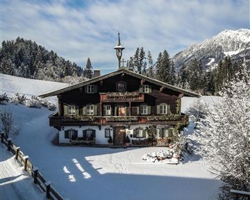 Ein traditionelles Chalet im Schnee, umgeben von Tannenbäumen. Der Himmel ist blau und wolkig, was eine idyllische Winterlandschaft schafft.
