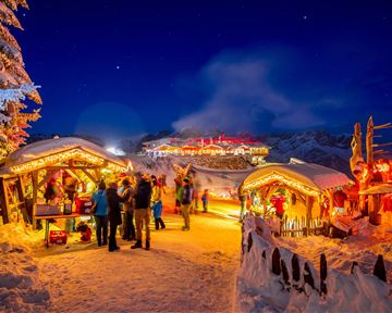 Ein festlicher Weihnachtsmarkt in einer verschneiten Landschaft. Die Stände sind mit Lichtern geschmückt und Menschen genießen die Atmosphäre bei Nacht.