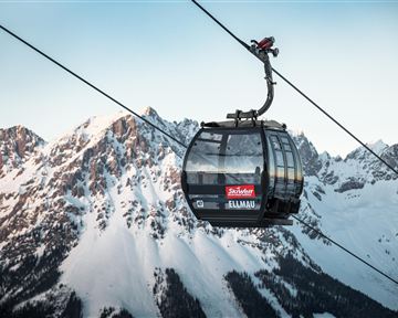 A cable car glides over snow-covered mountains. The sky is clear and the landscape appears majestic.