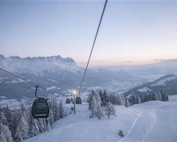 A picturesque winter landscape with snow-covered mountains and a cable car. The sky is clear with gentle colors of the sunset.