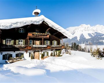 Ein charmantes Holzhaus im Schnee, umgeben von Bergen. Der klare blaue Himmel verleiht der Winterlandschaft eine lichte Stimmung.